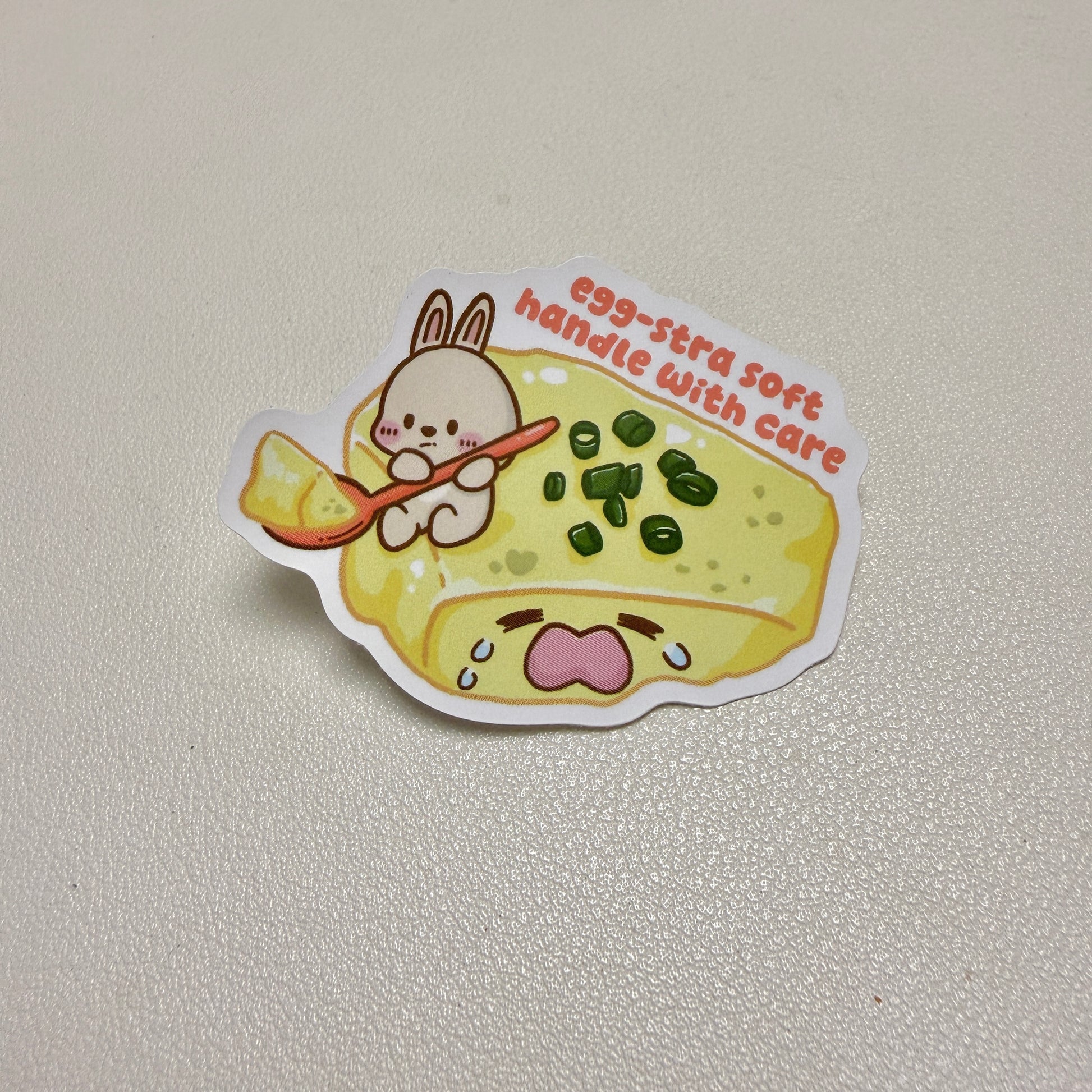 sticker of rabbit scooping a crying steamed egg with text 'egg-stra soft handle with care'