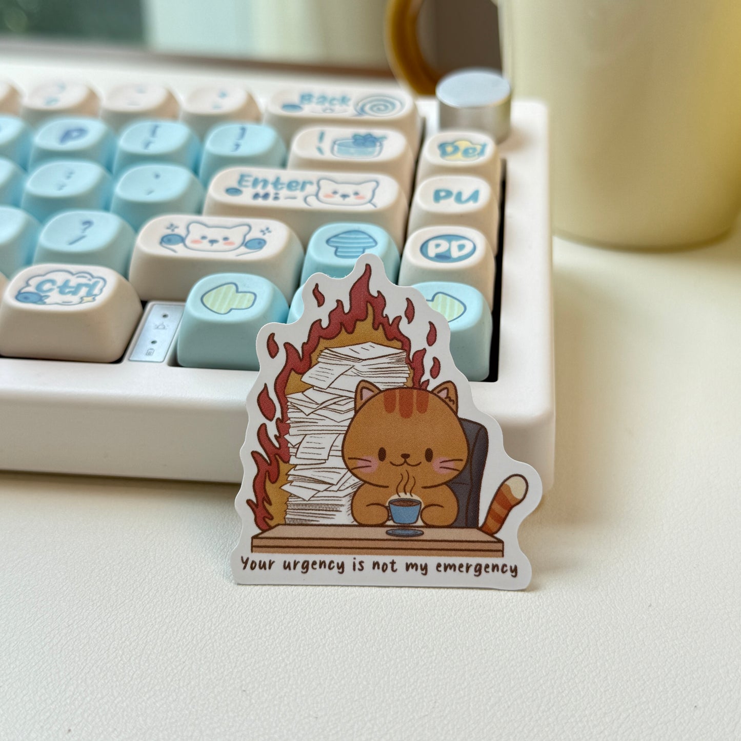 Sticker with a cat holding a cup of coffee and fire burning behind with text 'your urgency is not my emergency'